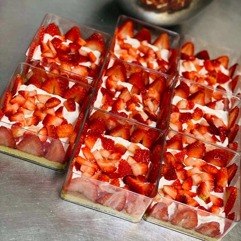 Nine clear containers of strawberry shortcake with whipped cream and fresh strawberries on a metal surface.