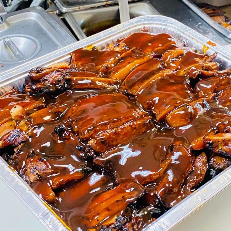 A large foil tray filled with glistening, dark brown Hawaiian BBQ chicken wings and drumettes.